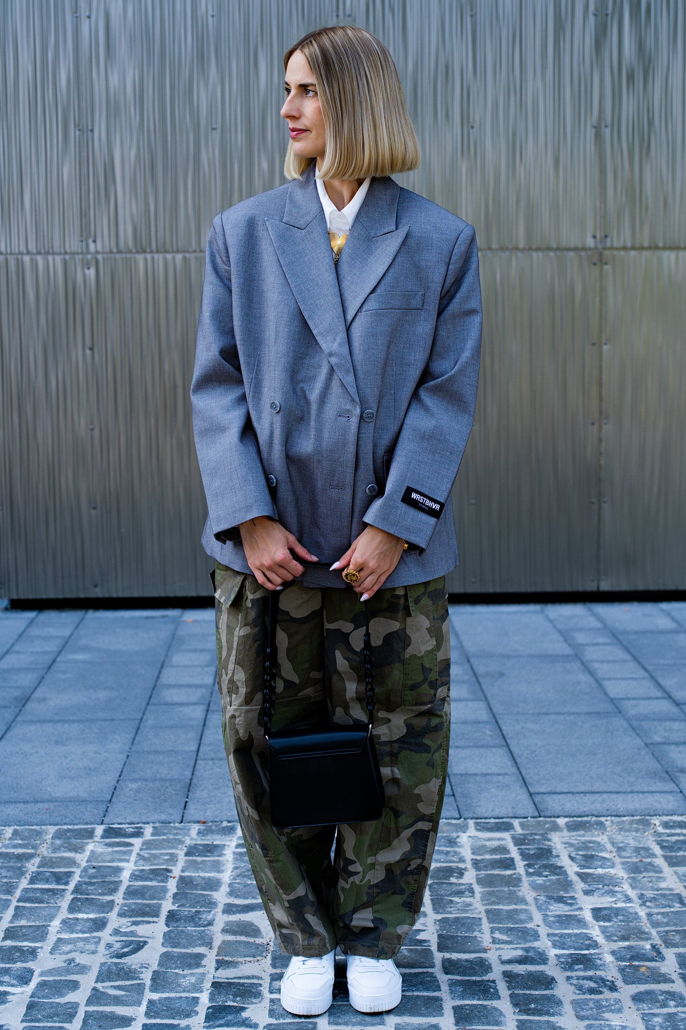 Frau mit blonden Haarn trägt Camouflage Baggy, grauen Oversize-Blazer und weiße Sneaker, dazu goldenen Schmuck als Highlight. HEARTCORE Kollektion von schmucktick. 