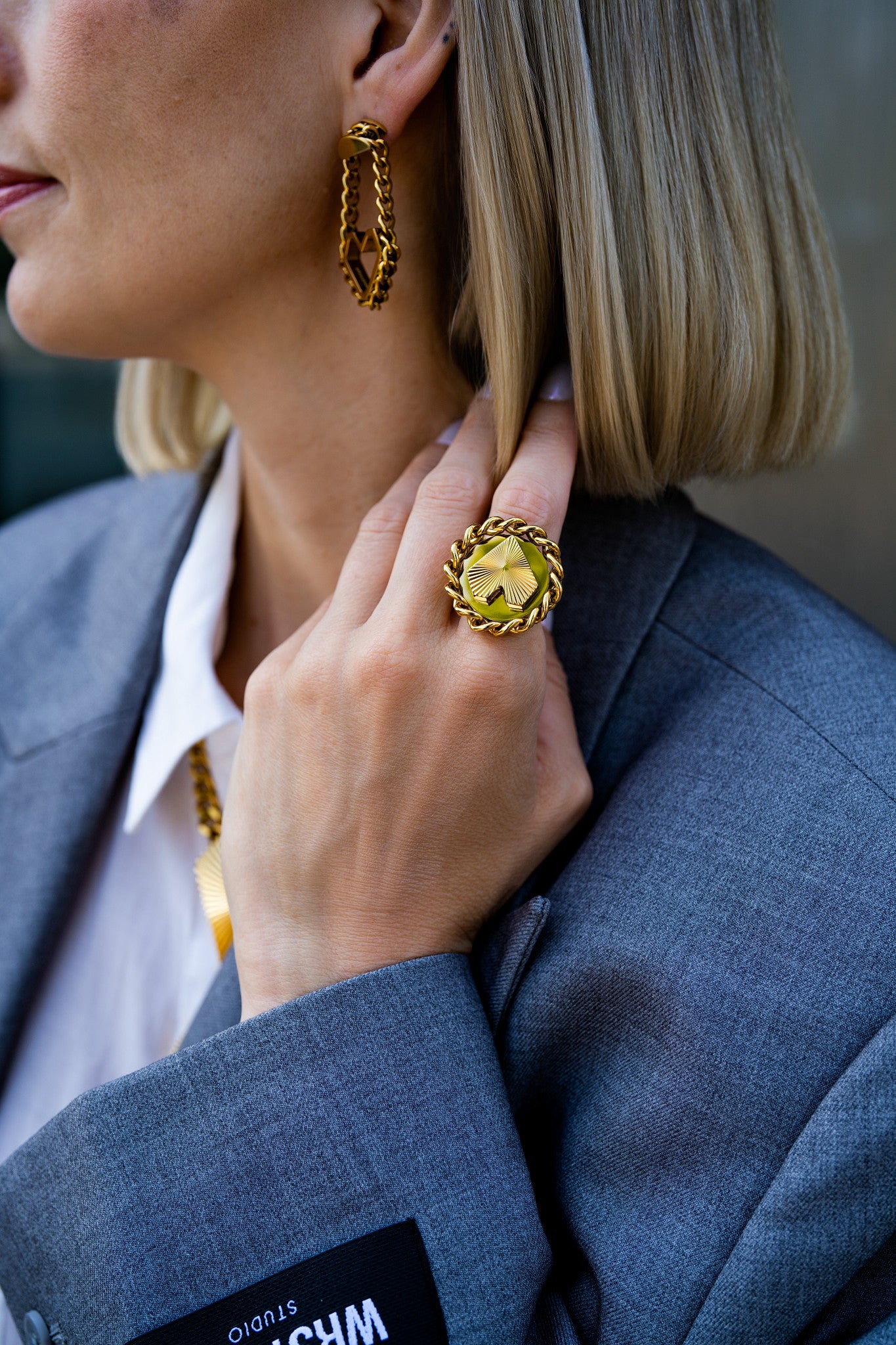 Nahaufnahme von goldenem Statement-Ring von Schmucktick mit Herz und Sunburst-Finish, Frau trägt grauen Blazer und Schmuck